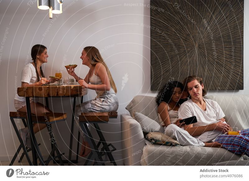 Cheerful women having dinner near flatmates using smartphone on sofa couple friend living room eat together cheerful gadget dine enjoy device couch browsing