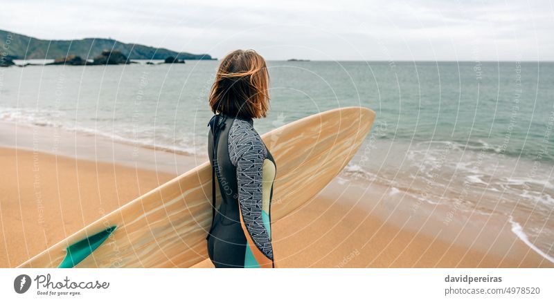 Surfer woman with wetsuit carrying surfboard looking to the sea at the beach unrecognizable young surfer horizon panorama panoramic banner web header winter