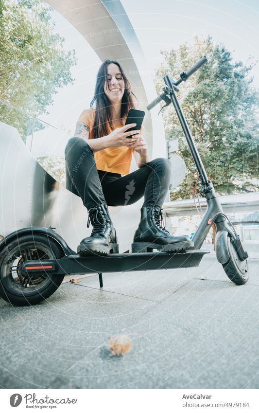 Portrait of young woman sitting on urban scenery holding a mobile phone and leaning on an electric scooter with a smile. Resting and watching social media with casual clothes. City lifestyle