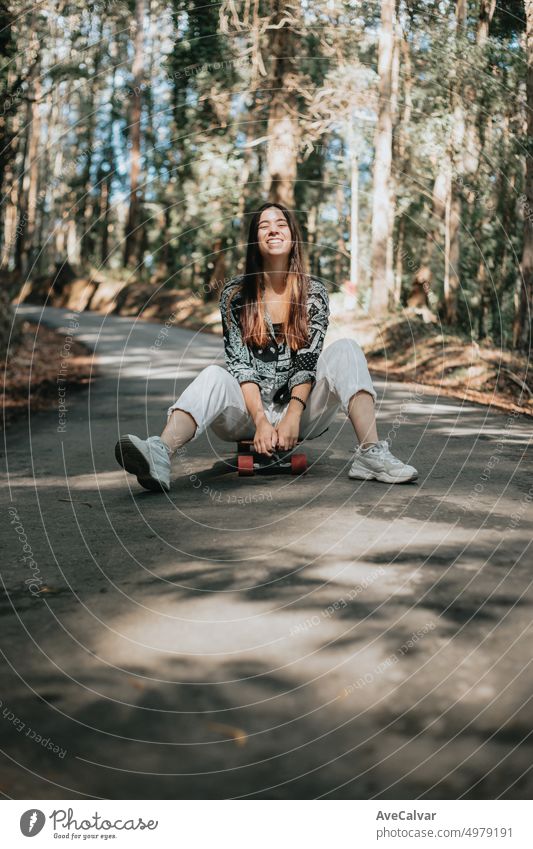 Young woman in a path in the middle of the road surrounded by forest sitting above a longboard skate. Excited and enthusiastic for new hobby.Resting after a long trip doing sightseeing.Freedom concept