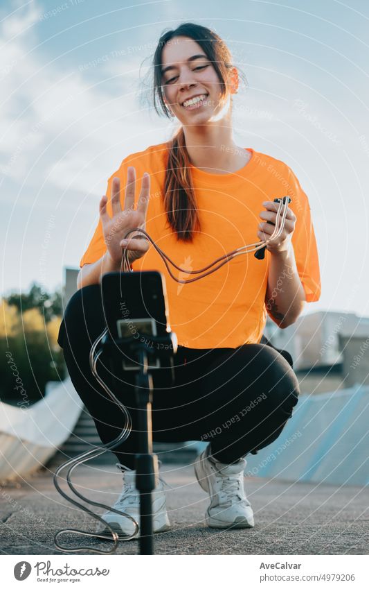 Young adult woman recording a live stream waving hand and holding a jump rope.Coach training, explaining,teaching and answering questions about calisthenics. Outdoor sports.Casual and sporty clothes