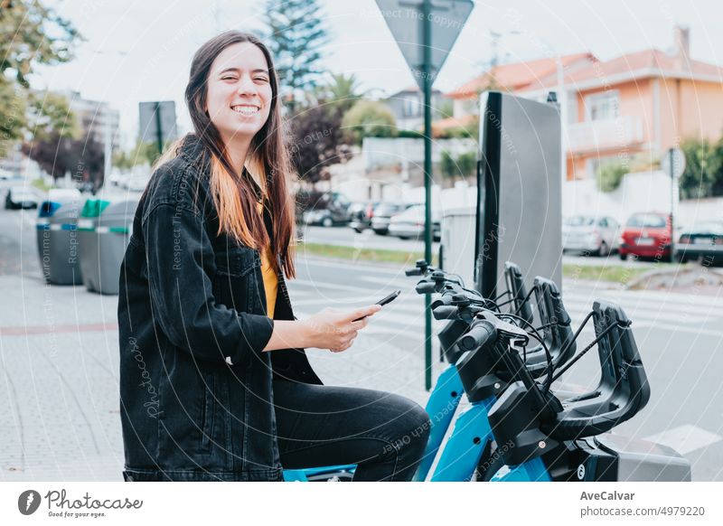 Young woman smiling checking her phone to unlock and rent a bike. Checking if there is any available bicycle and battery status. Electric transport concept. Renewable resources.Moving around the city