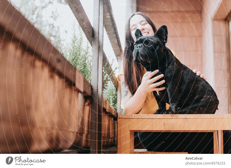 Young woman petting and hugging her black french bulldog while sitting on a wooden bench. Dog observing the city and girl smiling. New life in city, open and playful spot in terrace to play and relax