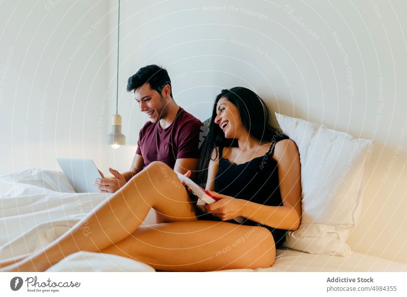 Couple with book and laptop lying on bed in bedroom couple woman linen using volume reading young white blanket browsing device gadget together love male female