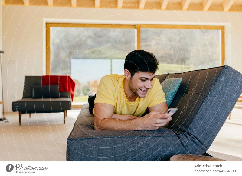 Young smiling man lying on sofa with smartphone in room hispanic guy settee mobile phone window using happy big young cozy home device gadget connection rest