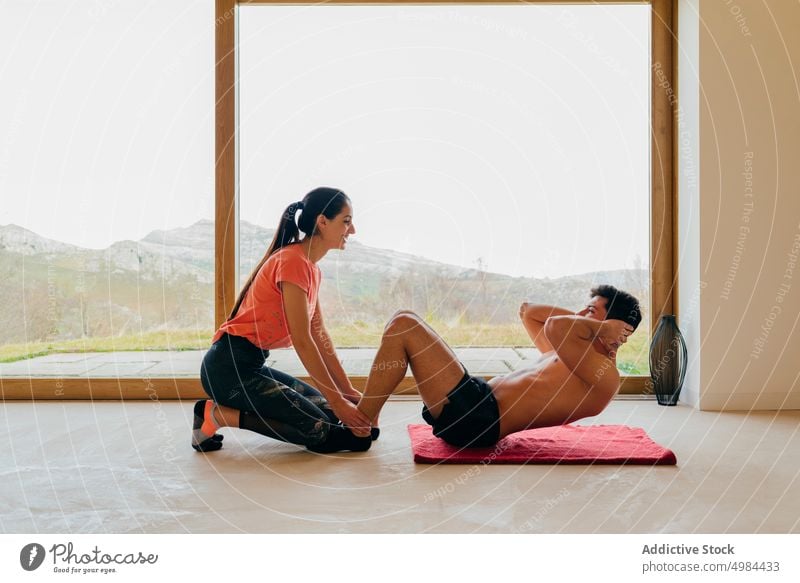 Happy woman helping man training in room near window couple sport sit up exercises shirtless leg happy young slim big sportswear lady guy athletic active