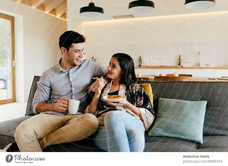 Happy couple with cup and plate on sofa in room happy hispanic kitchen mug spoon settee cheerful smiling woman young attractive beverage drink comfort meal