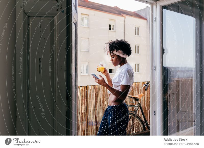 Healthy black woman browsing smartphone on balcony watching juice smile ethnic breakfast modern female morning drink young beverage cup device gadget memory