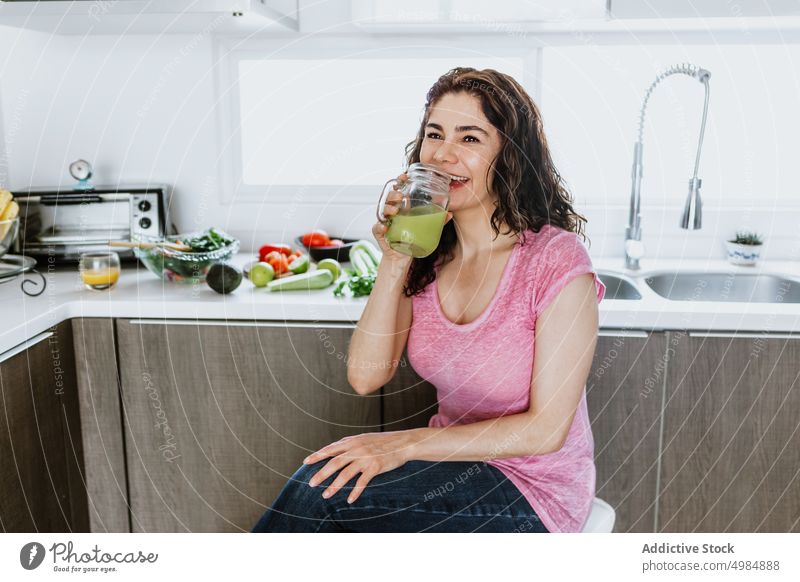Happy woman sitting in kitchen and drinking healthy green smoothie juice detox ingredient vegetable positive female cheerful toothy smile healthy lifestyle