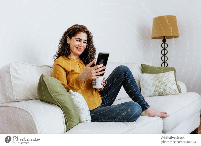 Smiling female talking via video call on smartphone at home woman smile positive using browsing cheerful cup coffee tea toothy smile curly hair interior lamp