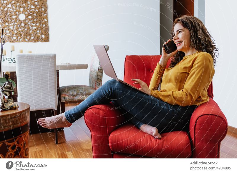Happy female talking on smartphone while browsing laptop in living room woman positive freelance phone call communicate cheerful gadget at home device using