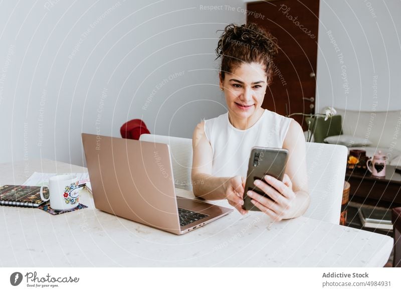 Smiling woman browsing on smartphone while sitting at table with laptop freelance positive video call work occupation female entrepreneur smile cheerful gadget