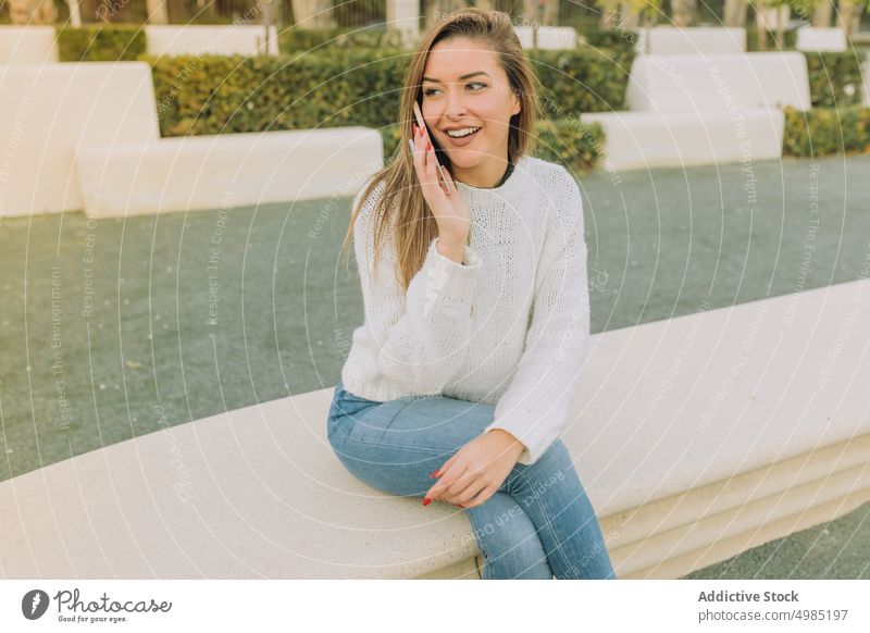 Delighted woman talking on phone sitting on bench phone call smile park cheerful happy female smartphone gadget device young sweater jeans conversation calm