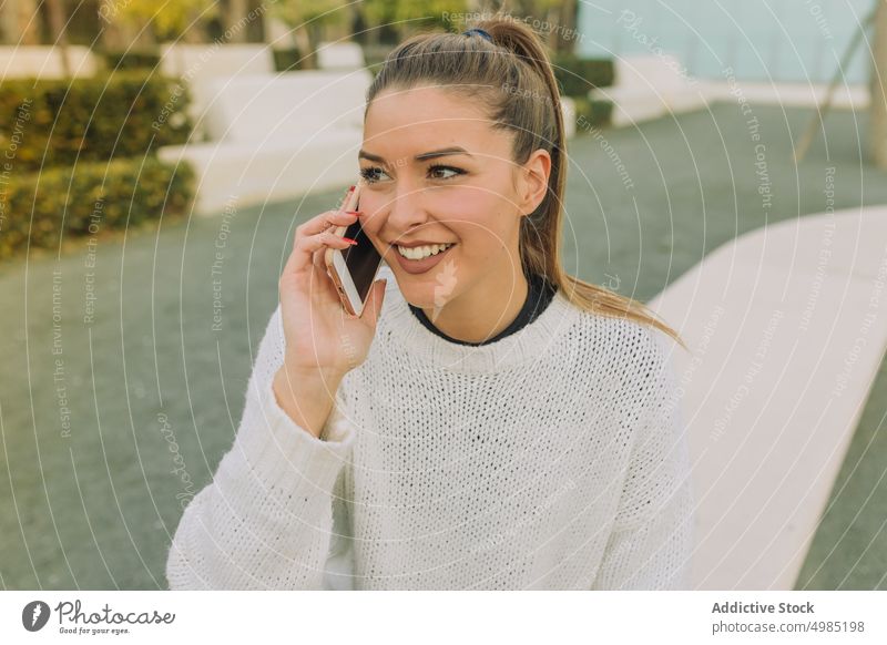 Delighted woman talking on phone sitting on bench phone call smile park cheerful happy female smartphone gadget device young sweater jeans ponytail conversation