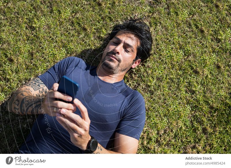 Tired ethnic sportsman browsing smartphone on green lawn tired break runner lying grass surfing online male hispanic grassy meadow internet athlete workout