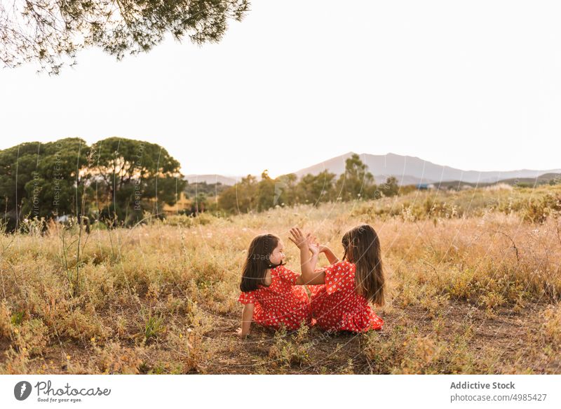 Two happy little sisters sitting in a meadow girl summer nature vacation carefree friend fun red playing family dress field sibling together childhood kid