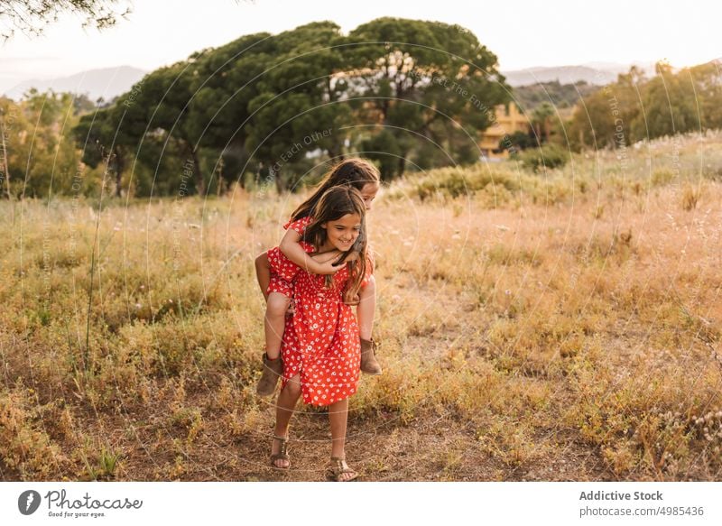 Smiling girl carrying sister on back in field piggyback summer nature ride vacation happy carefree friend fun meadow red walk family dress sibling together ears
