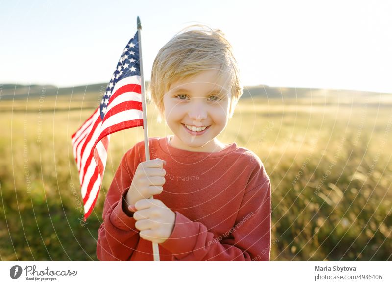 Cute little boy celebrating of July, 4 Independence Day of USA at sunny summer sunset. Happy child running and jumping with american flag symbol of United States over wheat field.