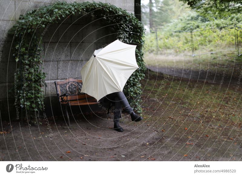 HH Tour Unnamed Street | A person sits on a bench and hides behind a large, bright umbrella. You can only see the legs. The bench is in front of a gray stone wall and is surrounded by an arch of ivy.