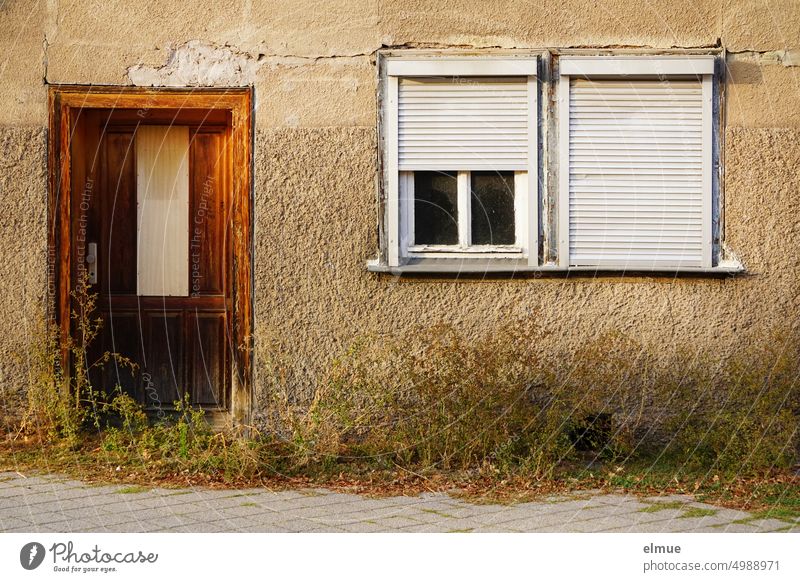 dilapidated residential house with old wooden door, two windows with half and full lowered blinds, respectively, and numerous weeds in front of them