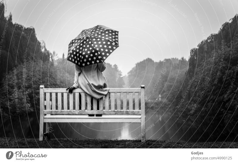 [HH Unnamed Road] Rear view of a woman in a light coat with a dotted umbrella. She is sitting on the back of a bench at the edge of a lake. Woman sedentary
