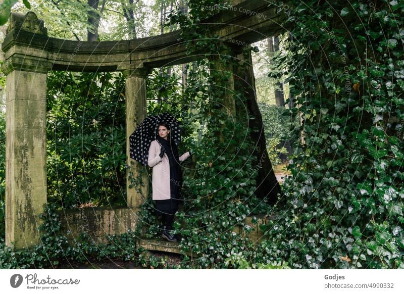 [HH Unnamed Road] Young woman with umbrella stands in arch of columns overgrown with ivy Woman Leaf canopy Tomb Column Monument Historic Colour photo