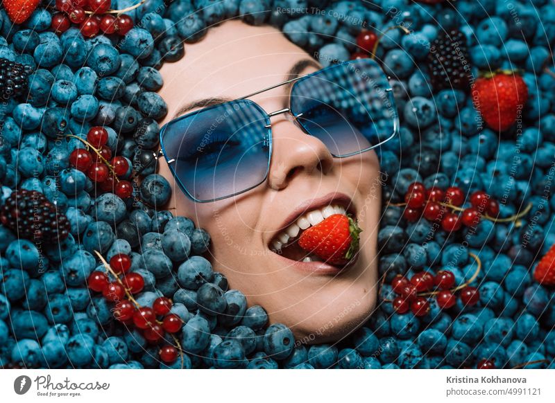 Happy woman face in eyewear fresh ripe berries - blueberries, strawberries, currant. Young girl covered with blackberry. Lady enjoying organic bilberry plant. Diet, antioxidant, healthy vegan food.