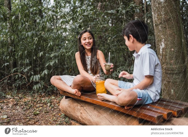 Smiling girl passing mate to brother on bench against trees drink beverage refreshment smile interact legs crossed cheerful herbal tea lemonade sibling