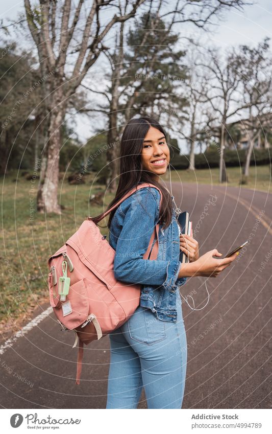 Young woman with backpack and smartphone on road student using smile park happy female young ethnic hispanic mobile cheerful positive denim jeans gadget style