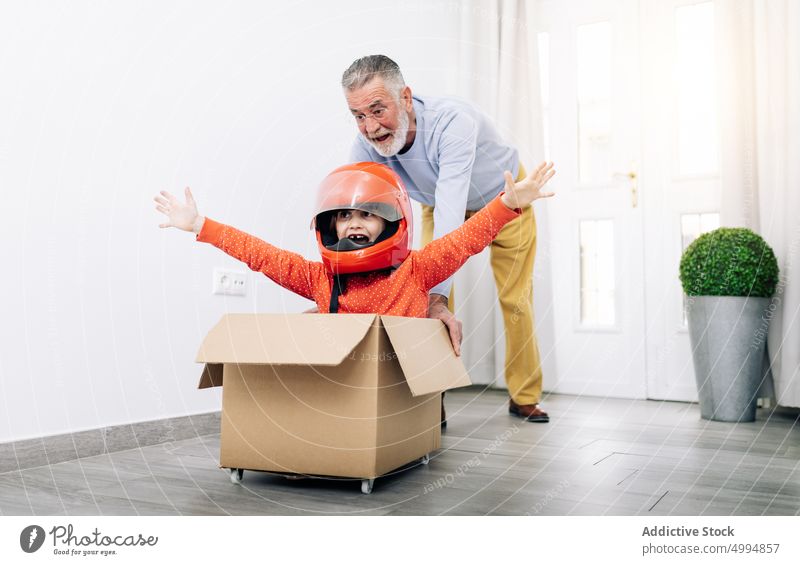 Child playing in racer in carton box with grandfather man kid dream pretend happy child playful win male middle age home helmet glasses light room door white