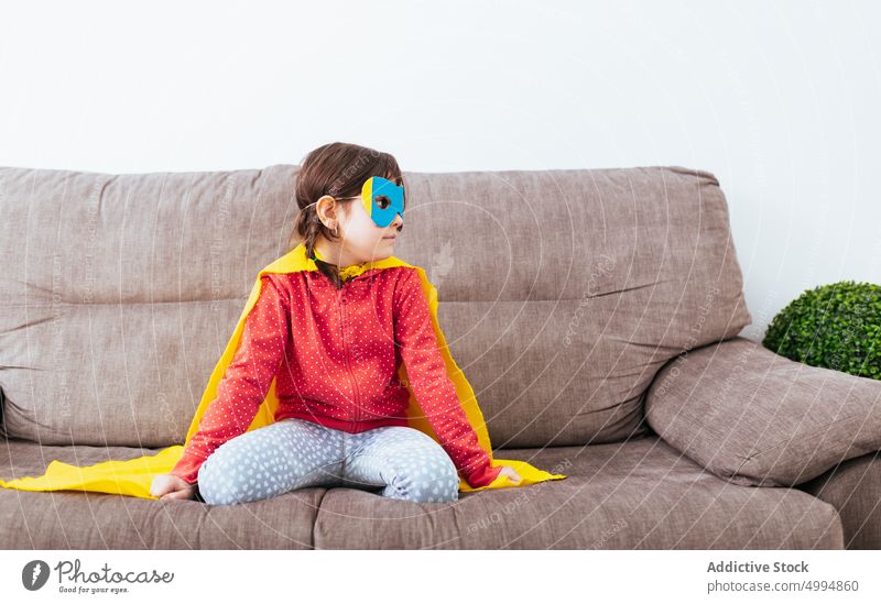 Girl in mask and cape pretending to be superhero girl costume confident childhood room kid sofa light home white wall little imagination couch courage fearless