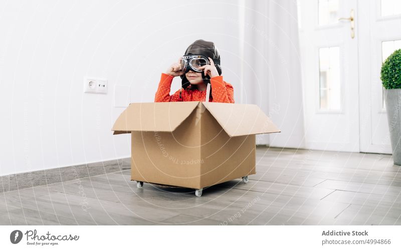 Child in pilot helmet and goggles dreaming about future kid aviator pretend dreamy imagination thoughtful childhood carton box light room white door potted