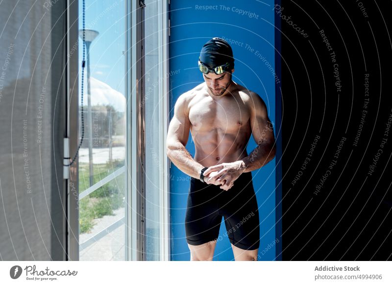 Muscular swimmer with naked torso near window looking at smartwatch athlete six pack abs dreamy macho masculine body man portrait smart watch brutal virile