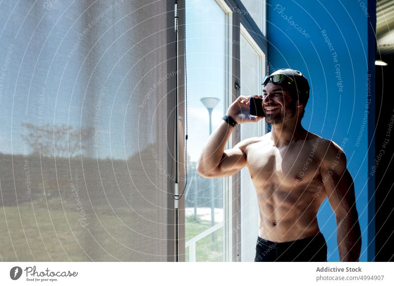 Cheerful swimmer talking on smartphone against window naked torso six pack muscular smile man