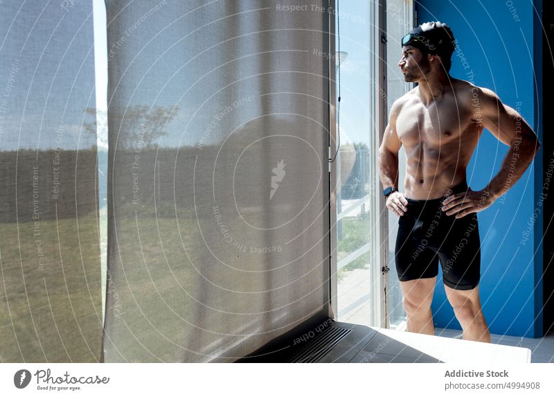 Muscular swimmer with naked torso behind window in sunlight athlete six pack abs dreamy macho masculine body man portrait ponder brutal virile manly abdomen