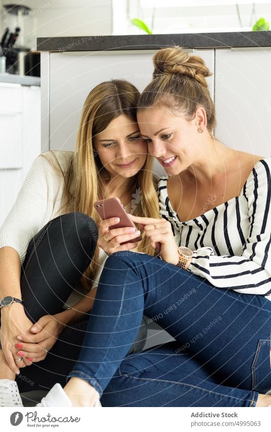 Young female friends sitting on floor in kitchen and sharing smartphone women together using free time social media show mobile young long hair casual browsing