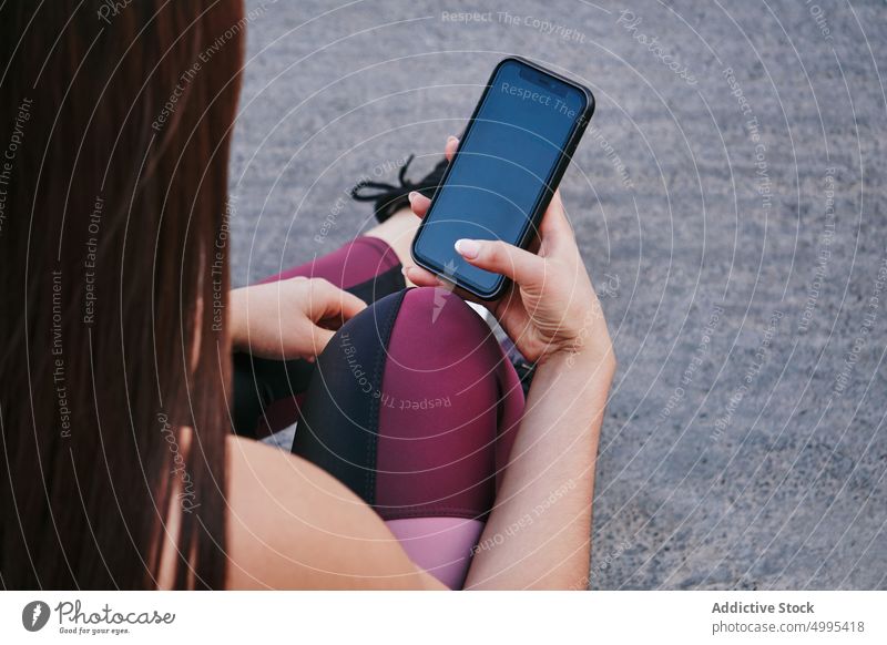 Crop sportswoman using smartphone on street break training fitness social media online ground female athlete sit mobile black screen device app digital