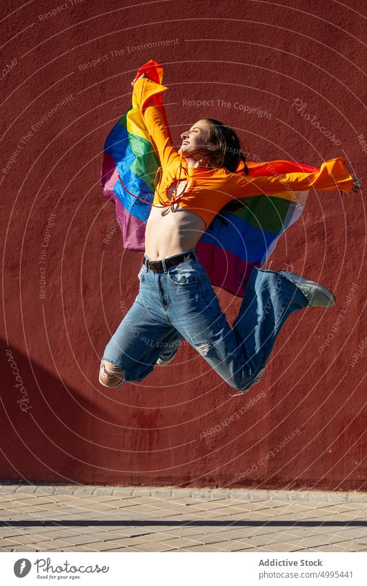 Young lesbian jumping with LGBT flag woman lgbt pride street happy wall freedom female young colorful smile arms raised rainbow urban cheerful building leap