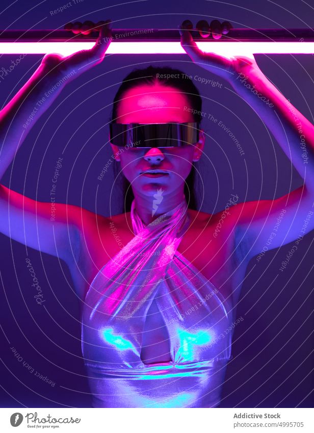 Futuristic young woman in trendy mirrored sunglasses with neon sticks in studio virtual reality futuristic self assured style confident portrait model