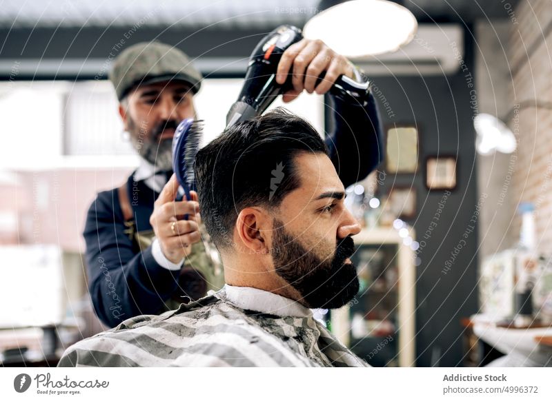 Middle aged ethnic barber drying hair of customer in barbershop men client serious hair dryer haircut comb salon concentrate beauty brutal confident mature