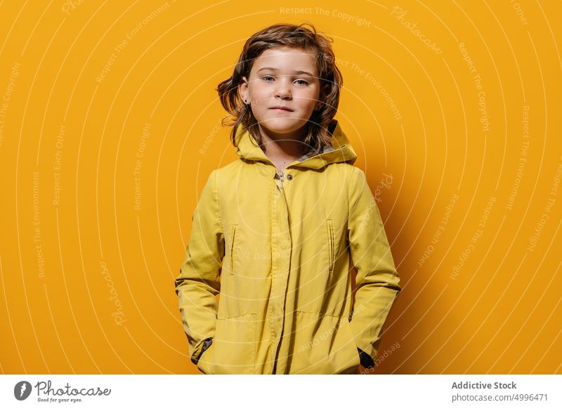 Girl looking at camera against yellow wall in studio girl colorful style autumn portrait childhood raincoat little vivid kid jacket winter trendy outfit joy