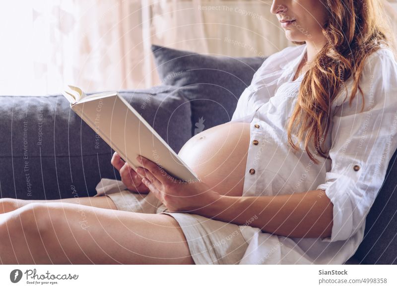 Pregnant woman reading a book at her home. white sofa living room sitting cozy bedroom morning sunday lying pregnancy pregnant female maternity mother happy