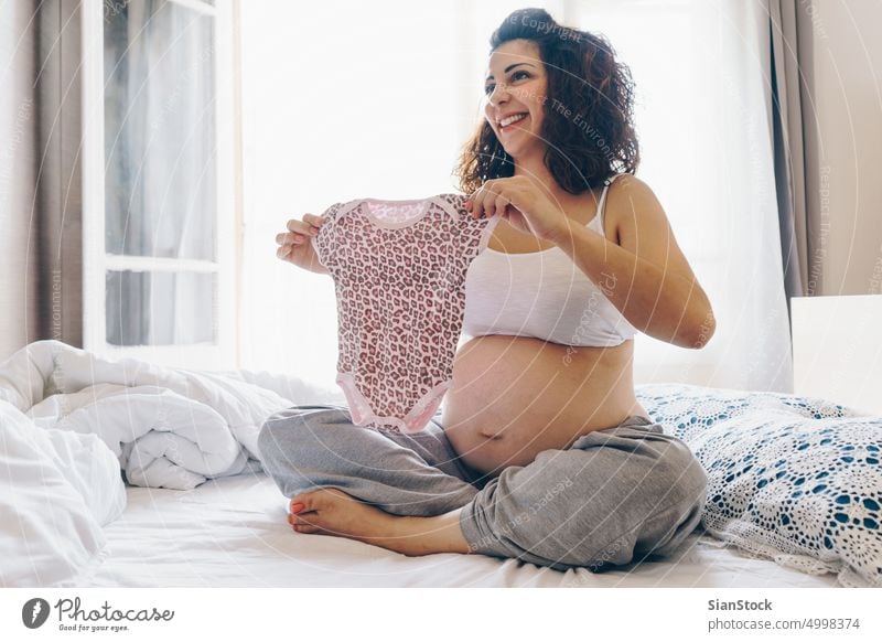 Happy pregnant woman holding baby clothing clothes pregnancy belly happy concept home beautiful mother natural light morning motherhood expecting bed bedroom