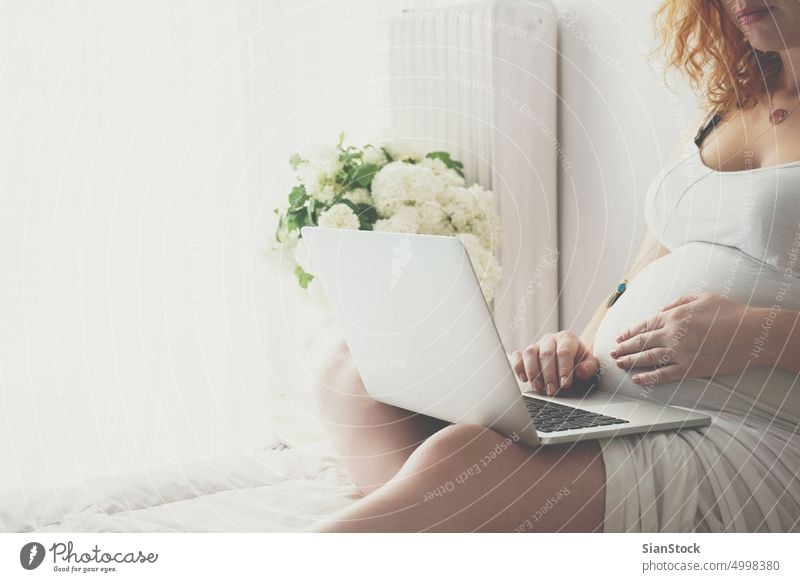 Young pregnant woman relaxing on bed with a laptop pregnancy morning young girl freckles sunday flowers dress belly female redhead red hair home bedroom job