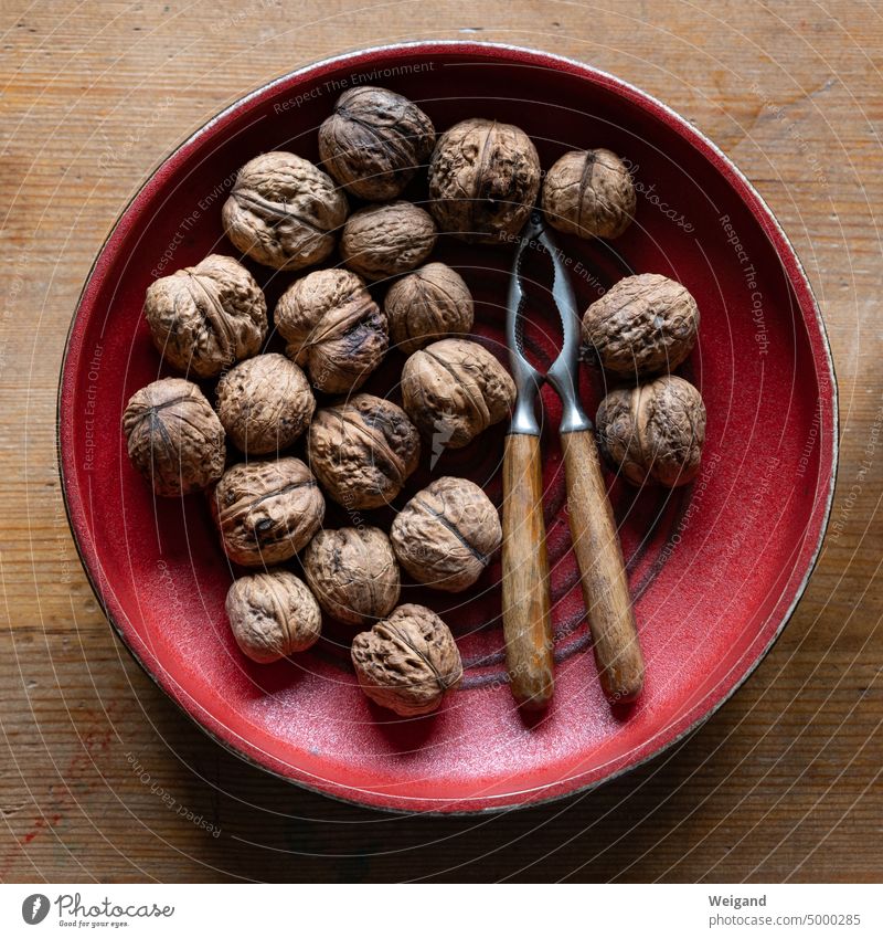 Round red ceramic bowl on a wooden table centrally photographed from above containing walnuts and a nutcracker with irregular wooden handle Central perspective