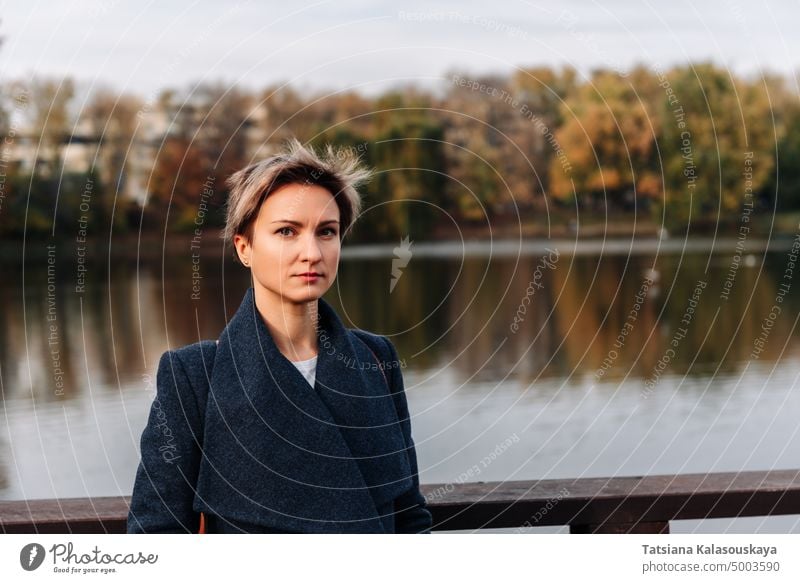 A young woman in a park in autumn by the river looks at camera fall short hair Blond Confidence female Lifestyles adult Leisure Independence middle aged