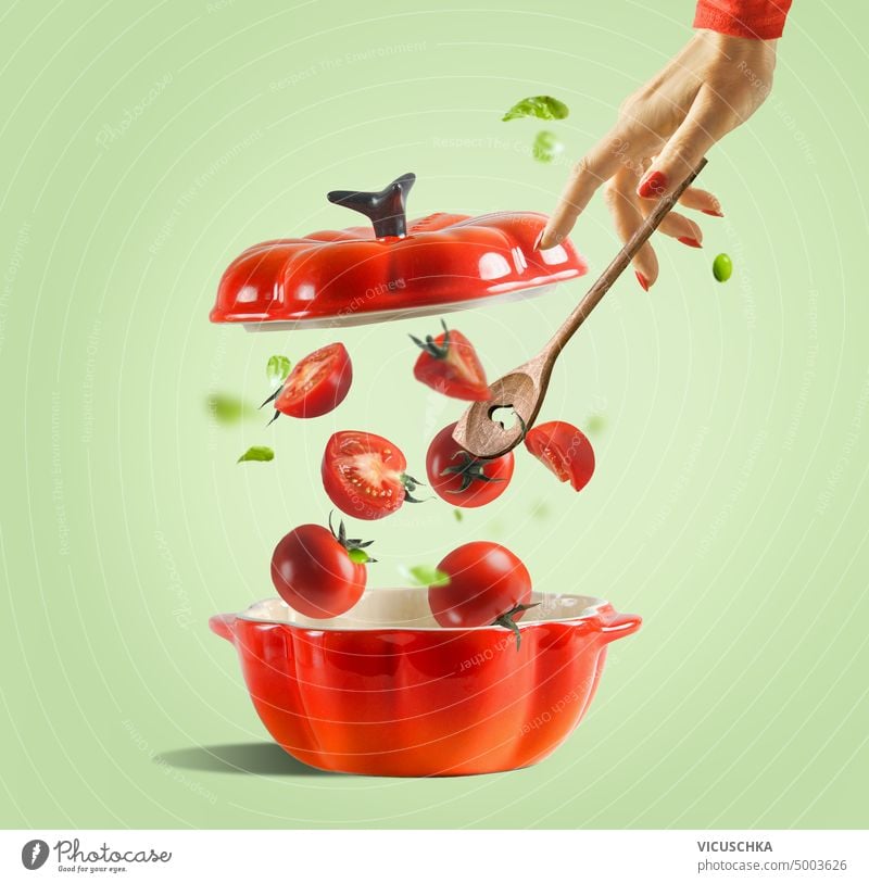 Preparation of tomato sauce or tomato soup. Red cooking pan with open lid and falling tomatoes and women hand with spoon on light green background. Food levitation.