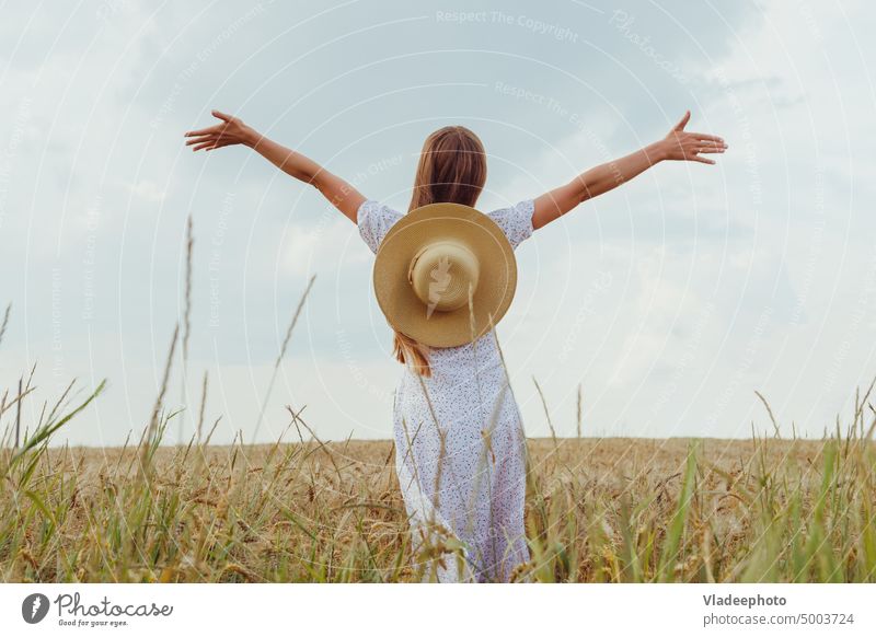 Young woman with hat raised hands up in wheat field back view. Concept of freedom nature summer rear female young beautiful girl happy dress sunny happiness