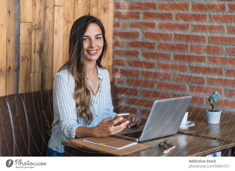 Smiling female freelancer using laptop in cafe businesswoman smartphone browsing social media smile project happy data remote telework table young smart casual