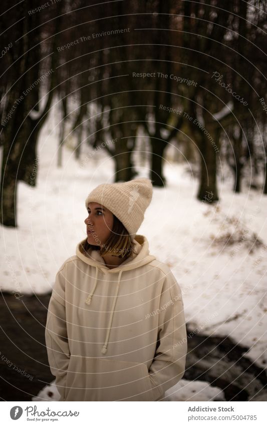 Young woman in warm clothes standing in winter woods snow cold dreamy river forest wintertime nature hat melancholy hand in pocket pensive calm woodland idyllic